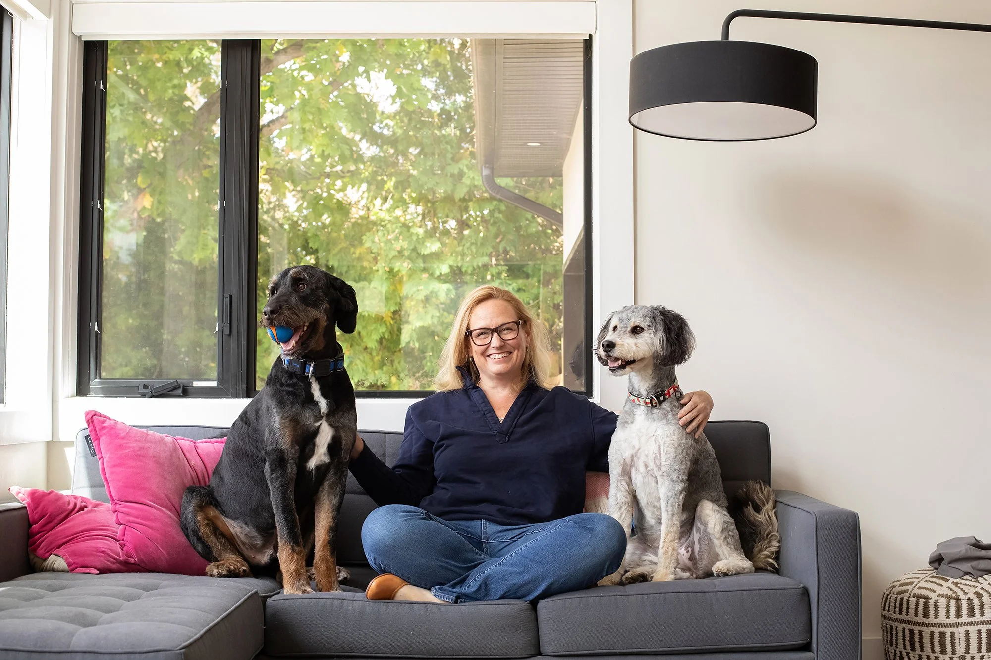 A woman sitting on a gray couch in a living room, holding two dogs, one on each side. The dog on the left is a large black and tan dog holding a blue ball in its mouth, and the dog on the right is a white and gray dog with curly fur. There is a pink pillow on the couch and a window with green trees outside in the background.