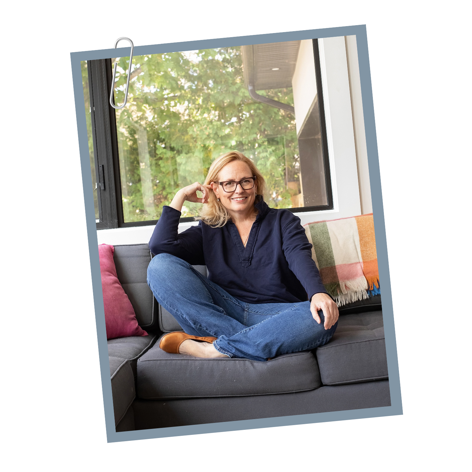 Woman sitting on a gray sofa near a large window with trees outside, wearing glasses, a navy blue shirt, and jeans, smiling and comfortably posed.