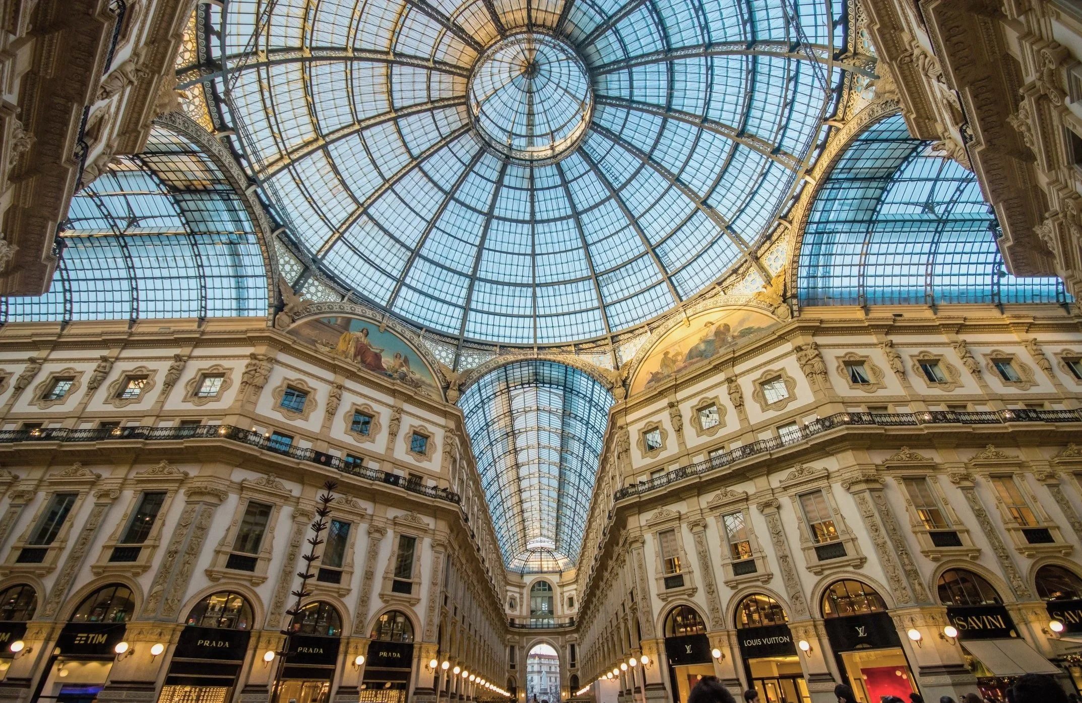The image shows a grand shopping mall with a large glass-domed ceiling. The interior features intricate architecture, multiple floors with balconies, and luxury brand storefronts like Prada, Louis Vuitton, and SAVINI.