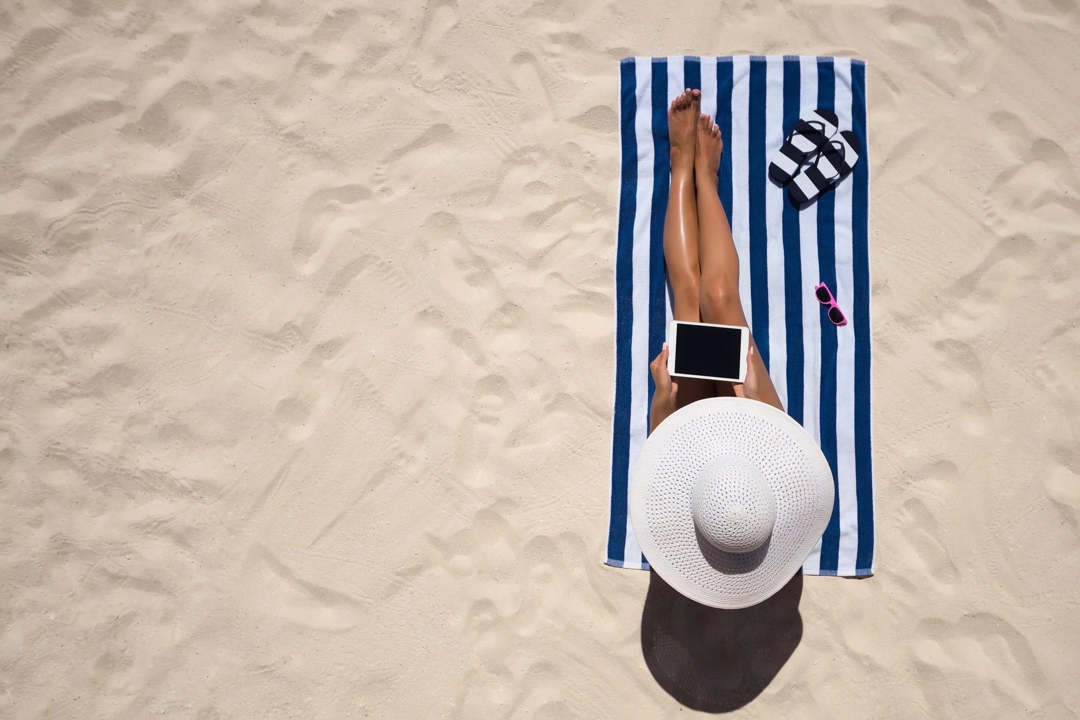 A person lying on a blue and white striped towel on the sand at the beach, holding a tablet with sunglasses and a wide-brimmed hat nearby.