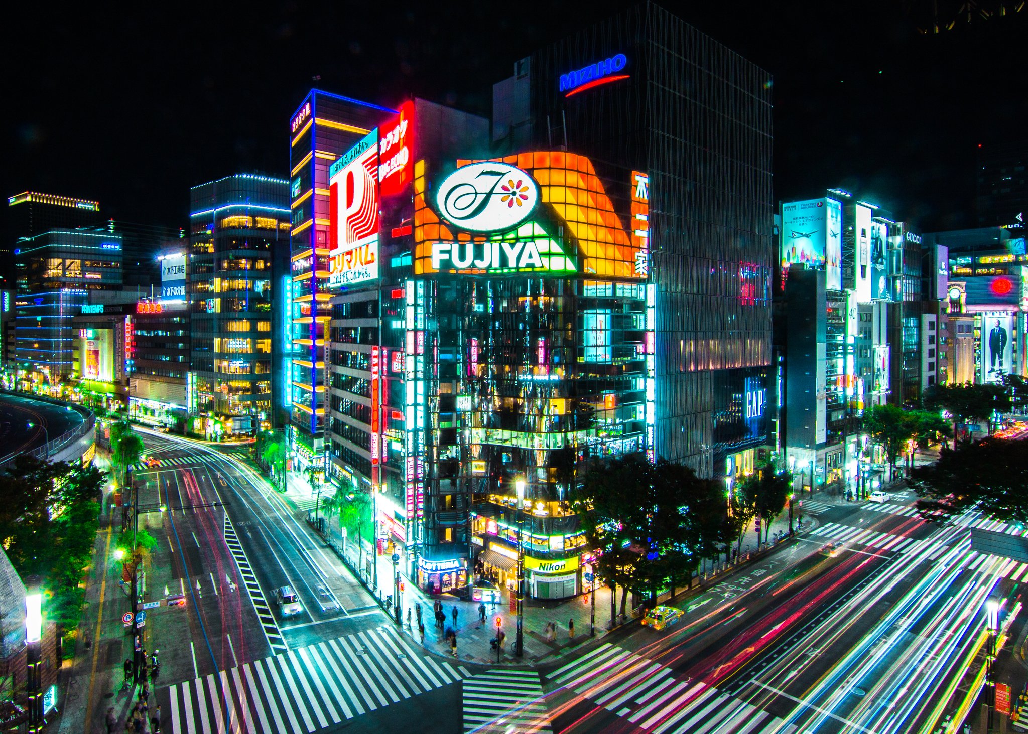 Nighttime cityscape with illuminated buildings, digital billboards, and streaks of car lights on the streets below.