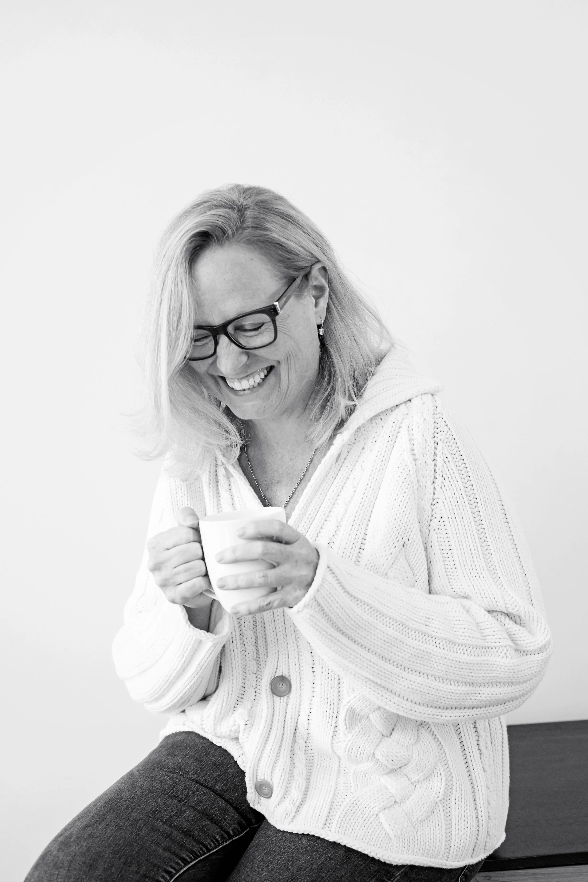 A woman with glasses and long hair, smiling, holding a mug, wearing a cardigan, sitting against a plain white background.