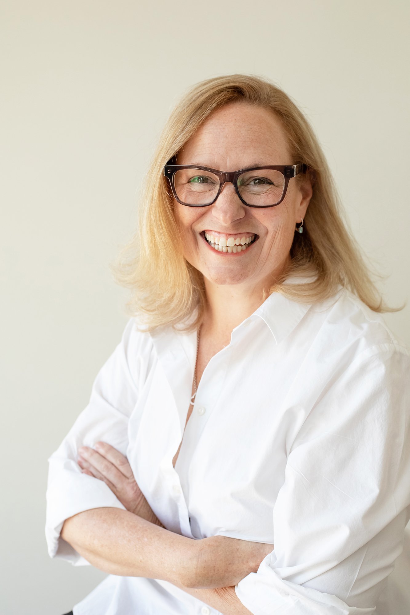 A woman with blond hair, glasses, and earrings smiling while wearing a white shirt, standing with crossed arms against a plain background.