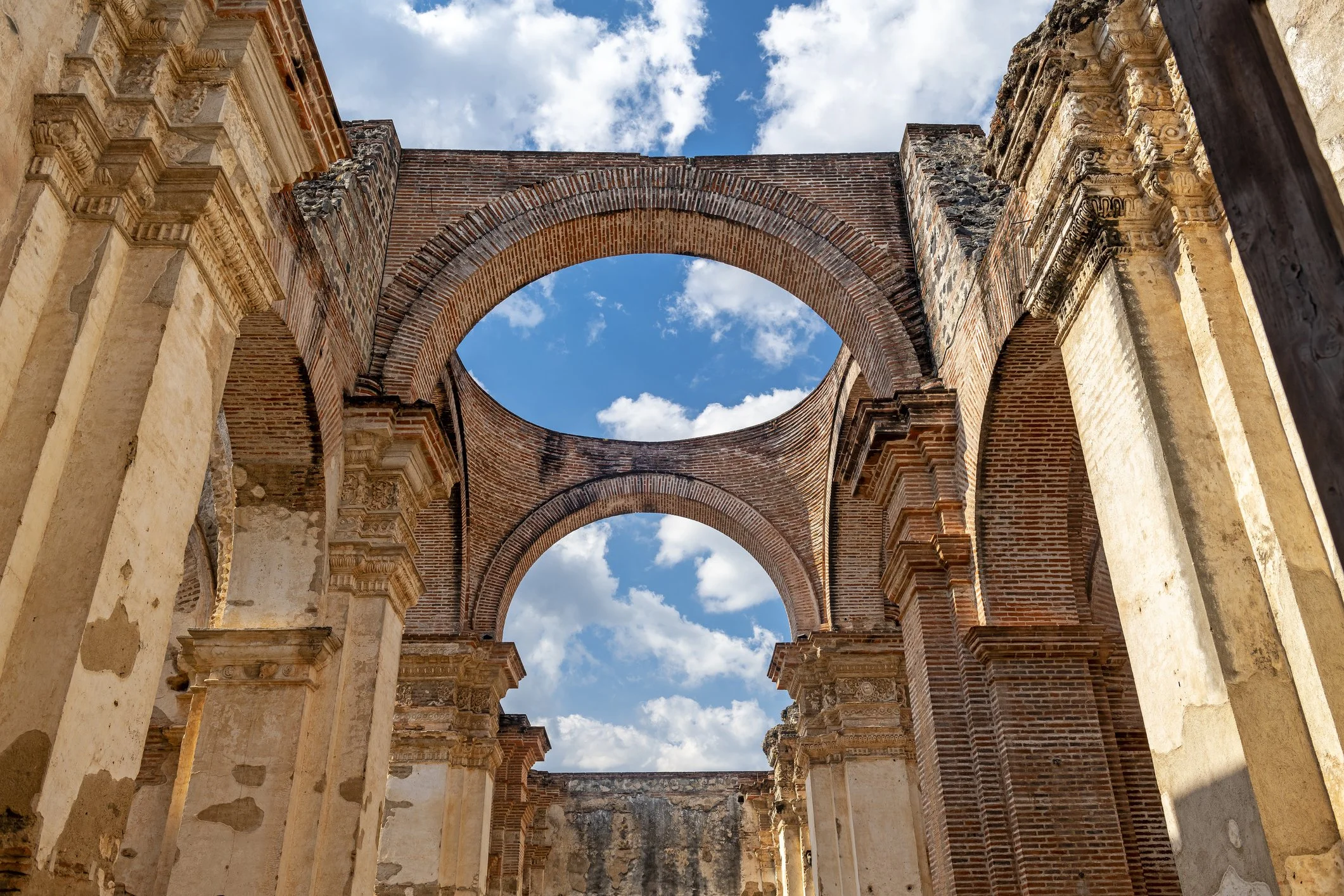 Ancient brick and stone ruins of a building with tall arches, set against a blue sky with scattered clouds.