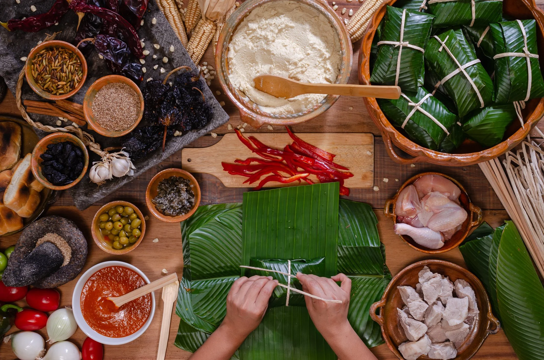 Person wrapping green banana leaves around food with a bamboo stick on a wooden table surrounded by various ingredients and dishes, including red chili peppers, garlic, a bowl of red sauce, and containers of rice, chicken, and vegetables.