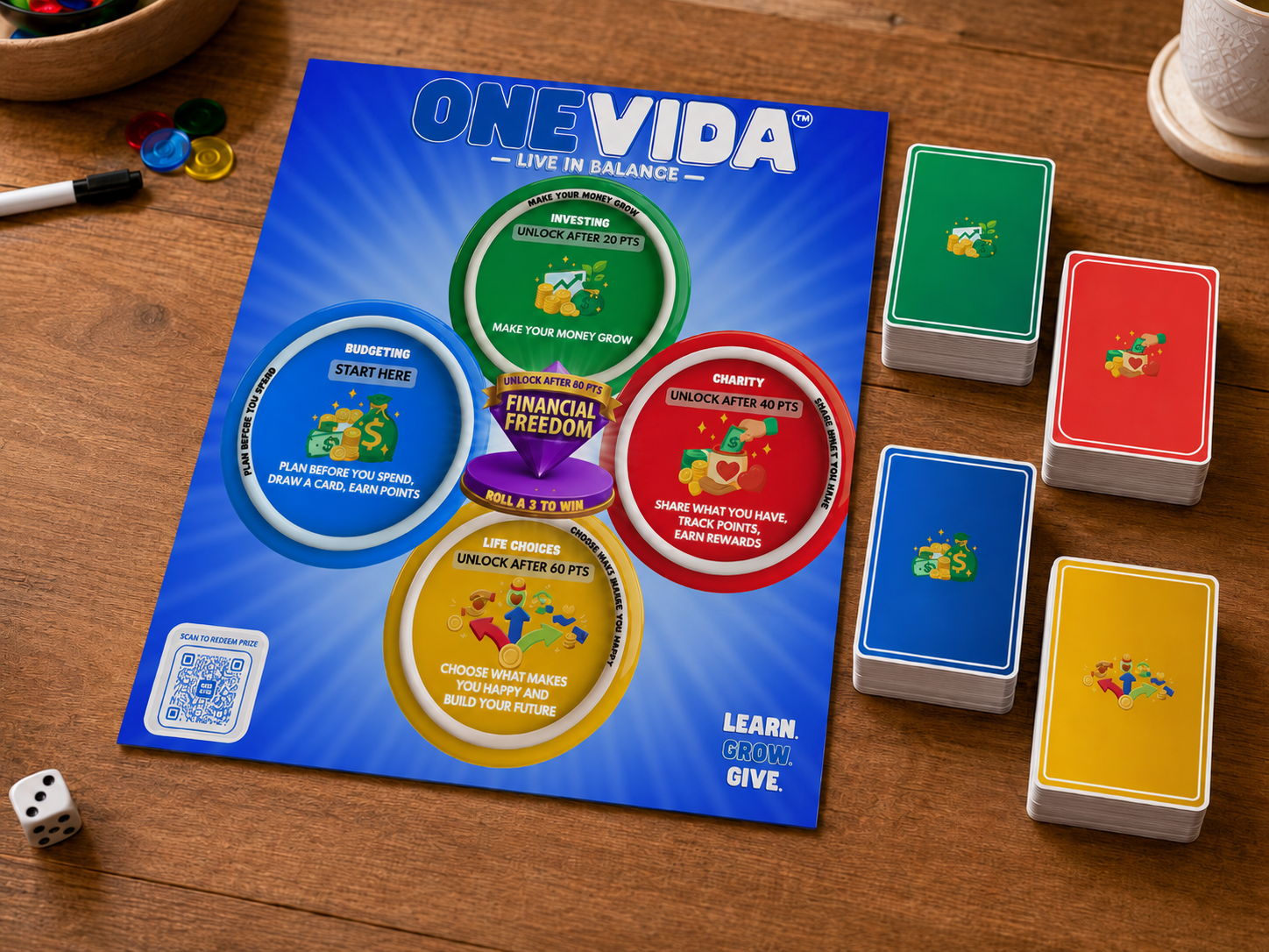 Board game called 'One Vida' with colorful stages for budgeting, investing, charity, and life choices. The game has stacks of cards in green, red, blue, and yellow. A die and small round tokens are also visible on a wooden table.