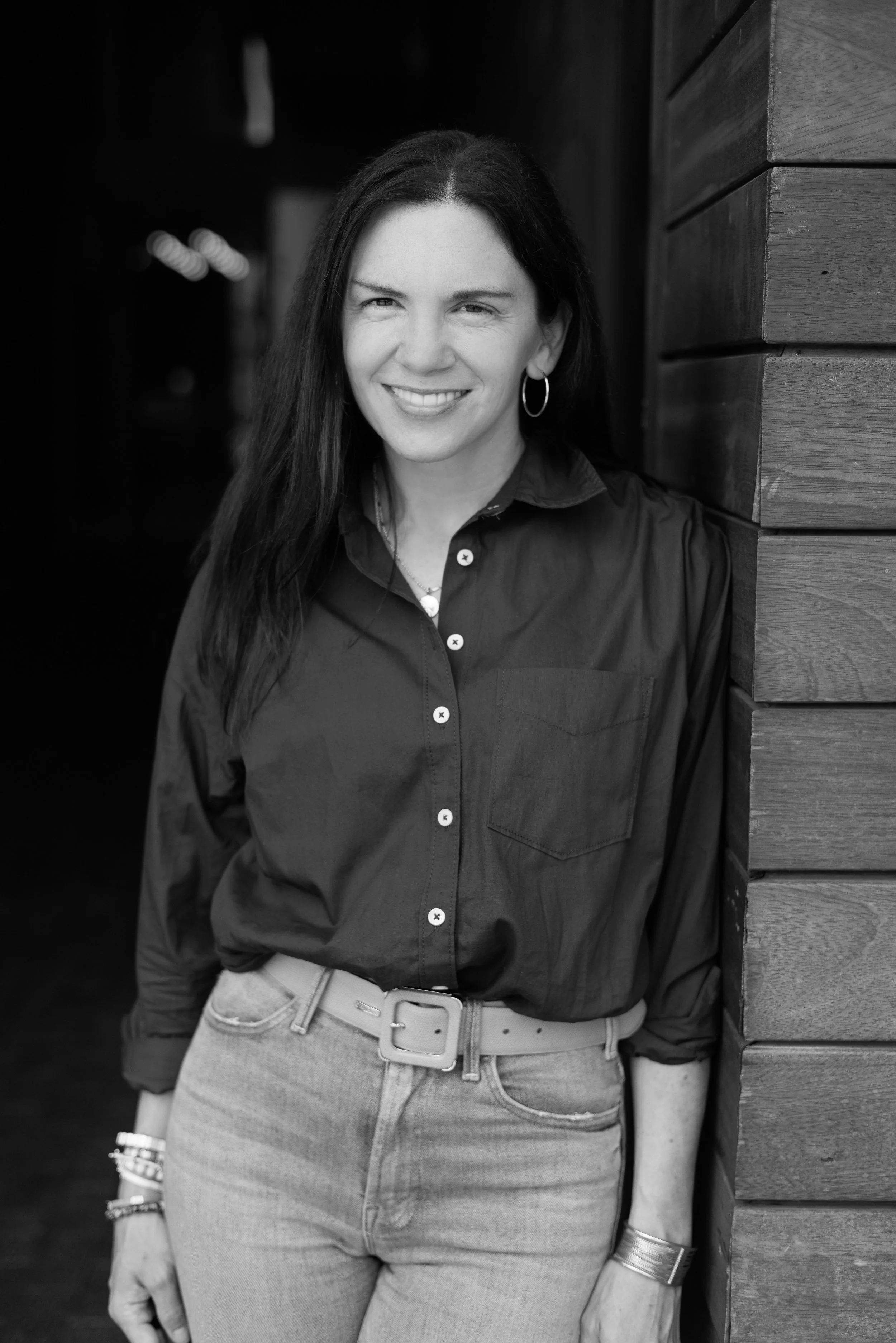 A woman with long dark hair, wearing a dark button-up shirt and light-colored pants, standing next to a wooden wall, smiling at the camera.