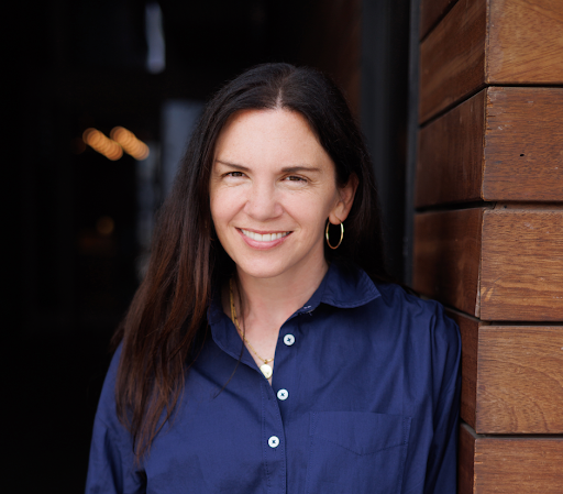 Woman with long dark hair smiling, wearing a blue shirt and gold hoop earrings, standing beside a wooden wall.