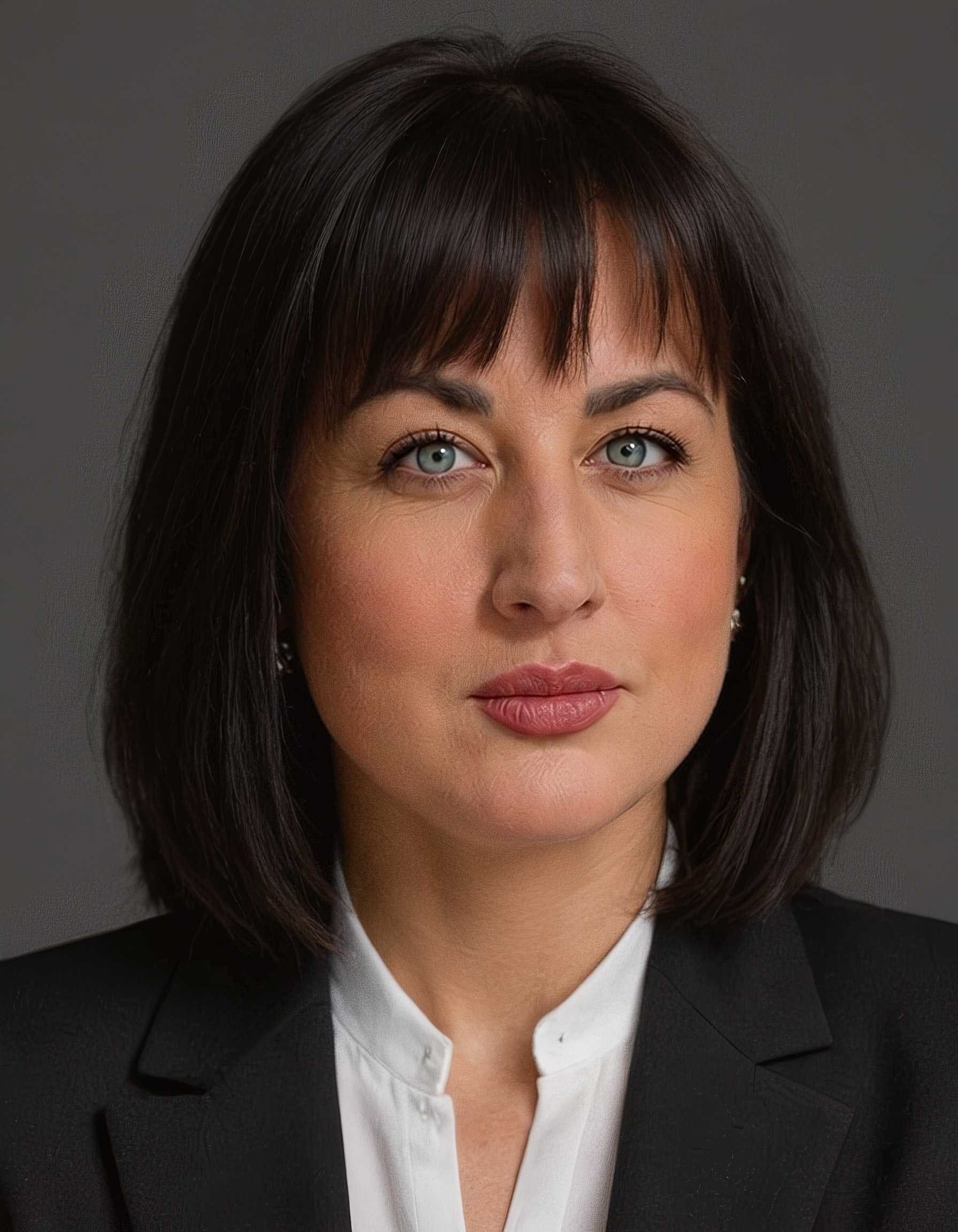 Close-up of a woman with dark brown hair, blue eyes, wearing a black blazer and white shirt, against a grey background.