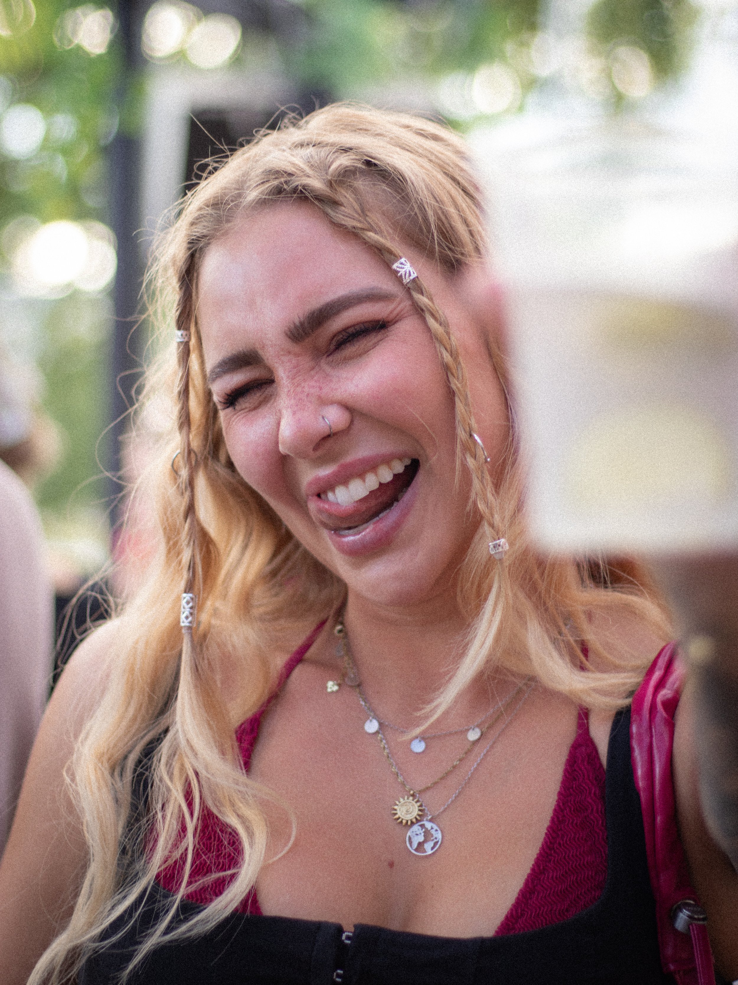 Eine lachende junge Frau mit blonden, gewellten Haaren, die mit kleinen Schmuckstücken frisiert sind, trägt mehrere silberne Halsketten mit Anhängern, ein pinkfarbenes Kleid und ein schwarzes Oberteil.