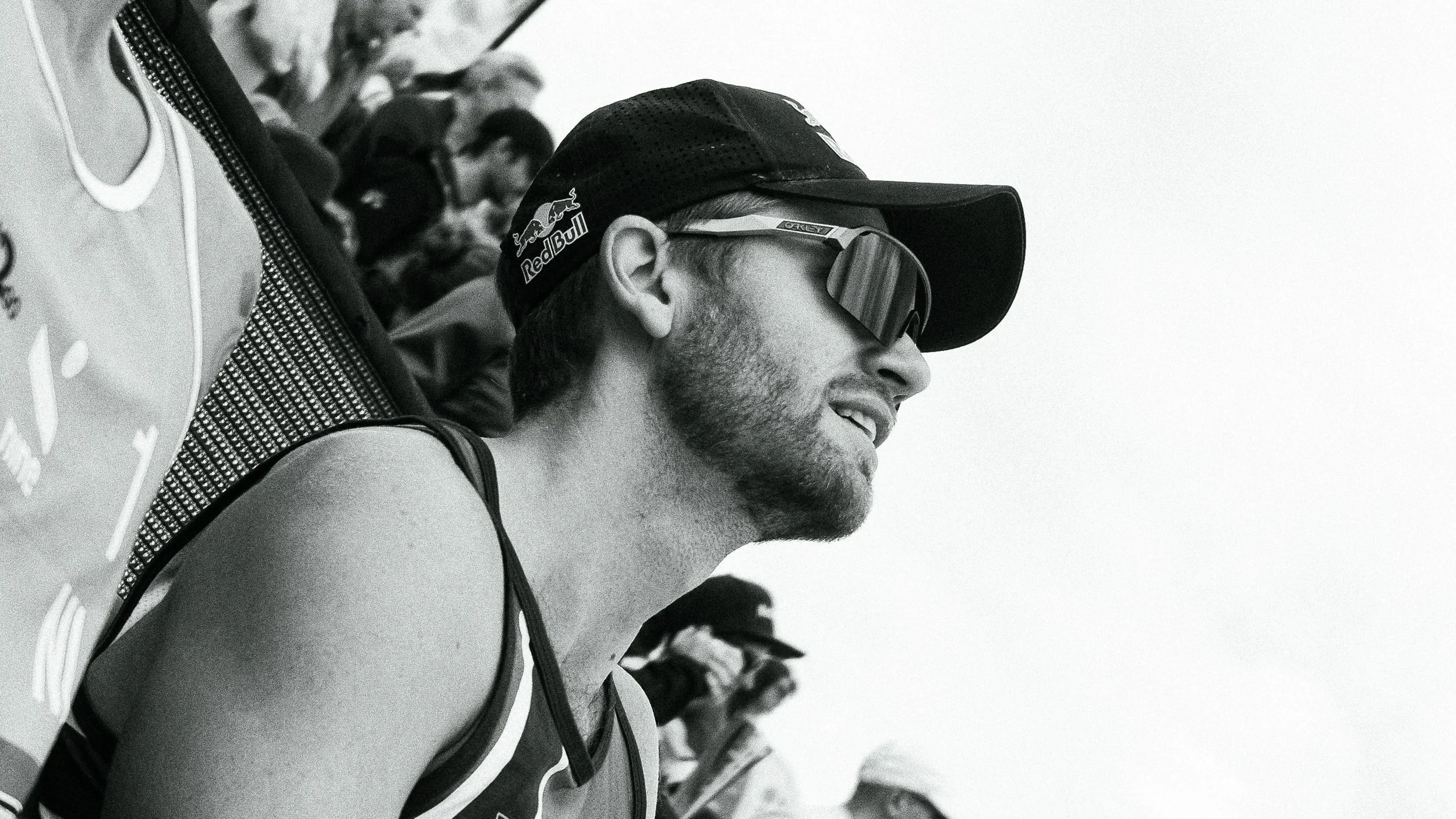 Ein junger Mann trägt eine Cap mit Red Bull Logo, Sportschutzausrüstung, und Sonnenbrille im Profil, im Hintergrund sind weitere Menschen zu sehen.