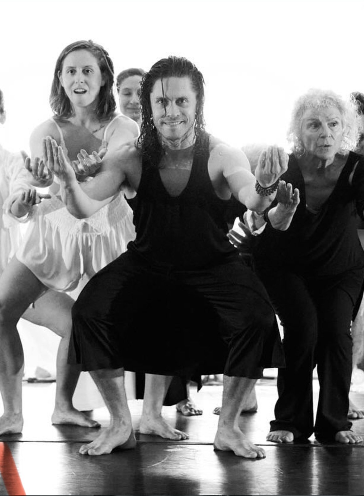 Grupo de personas participando en una clase de yoga o danza, en posición de cuclillas con los brazos extendidos hacia adelante, en un entorno interior con luz brillante.