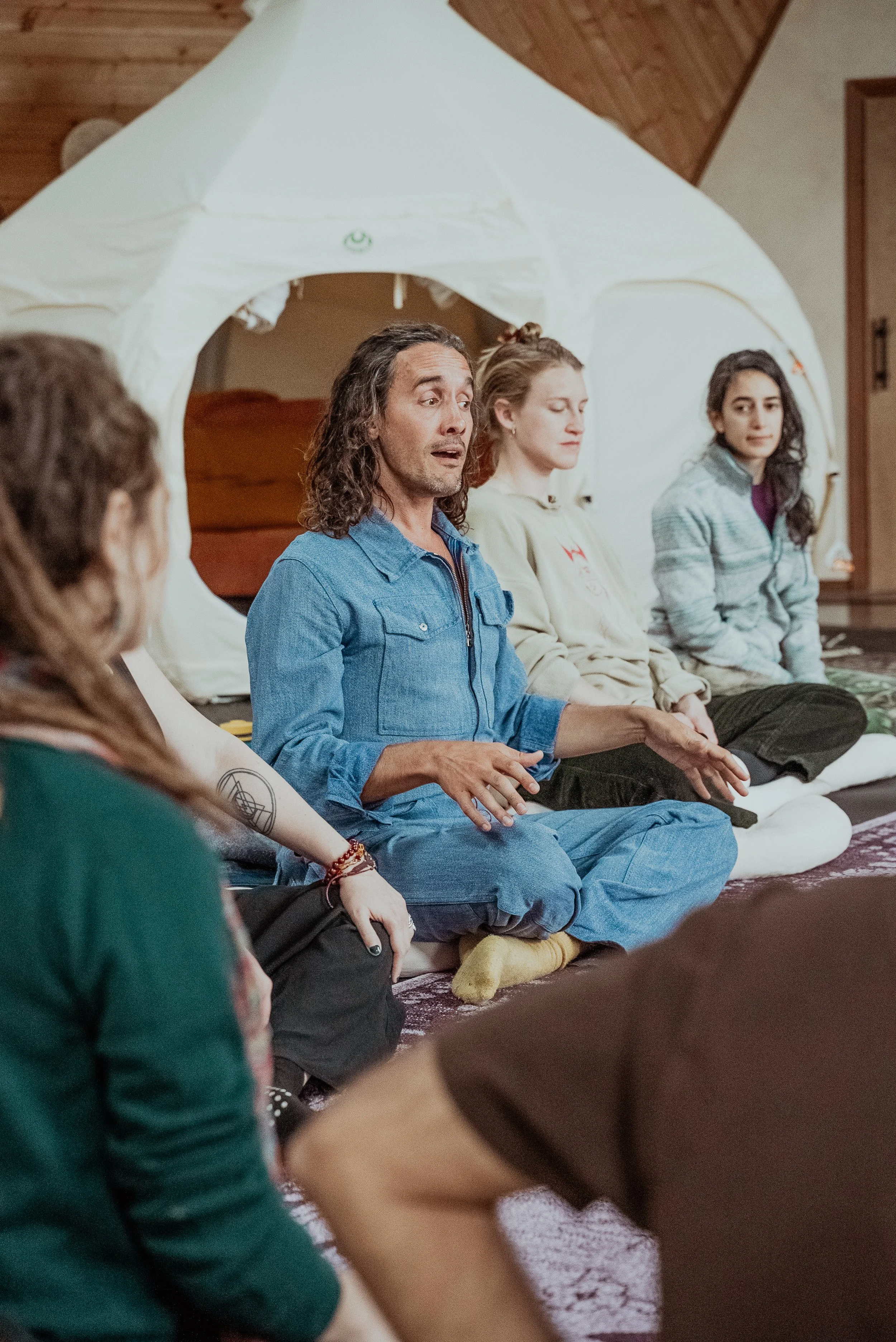 Un grupo de personas sentadas en círculo, participando en una sesión o reunión en un ambiente acogedor con techo de madera y una tienda tipi en el fondo.