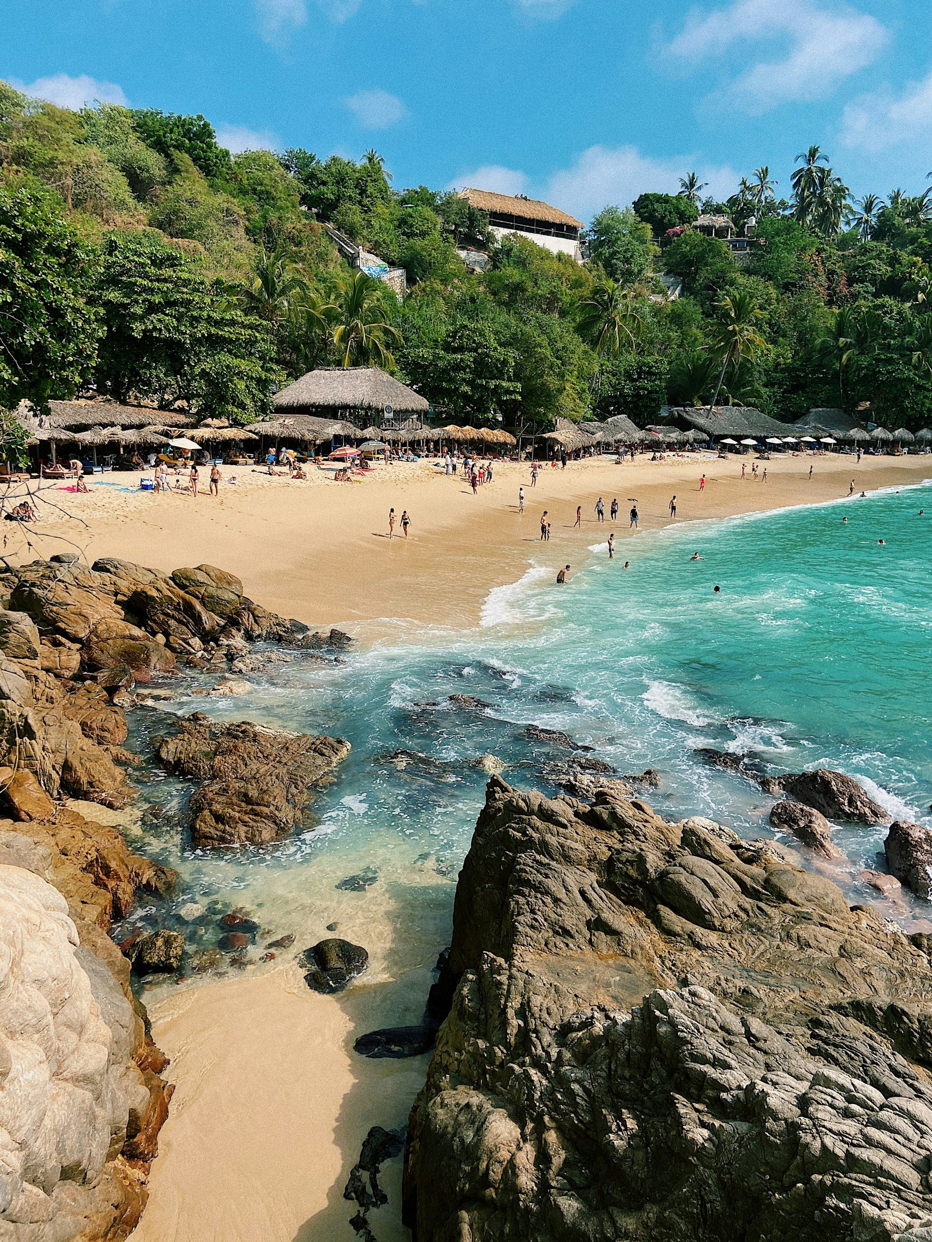 Playa con arena dorada, agua azul y rocas en primer plano, palmeras y casas en colina cubierta de vegetación en el fondo, cielo despejado con algunas nubes.