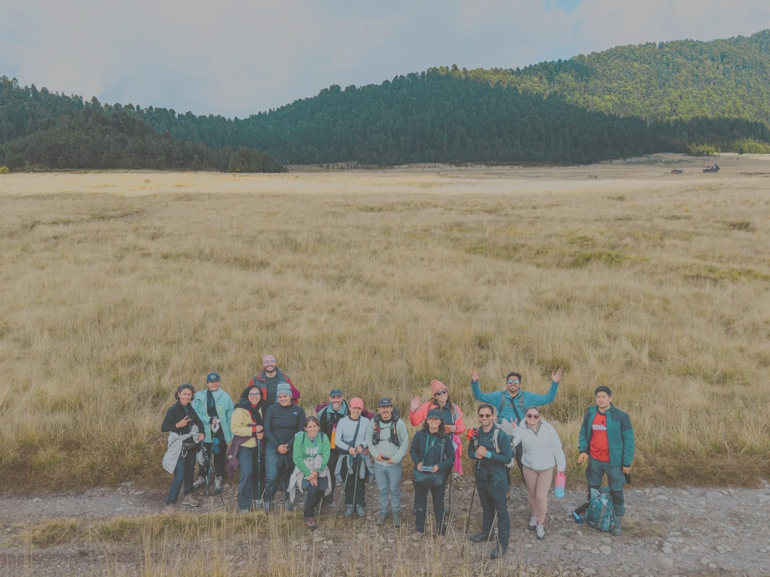 Grupo de personas con ropa de senderismo y mochilas, posando en un campo con colinas verdes y árboles al fondo, en un día nublado.
