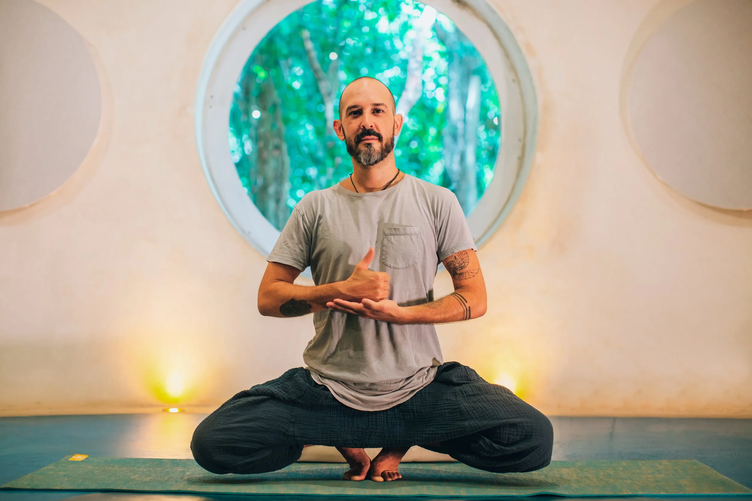 Un hombre con barba, sin cabello y tatuajes, sentado en posición de loto en una clase de yoga, sosteniendo un gesto de paz con una mano, en un aula con una ventana redonda con vista a árboles verdes.