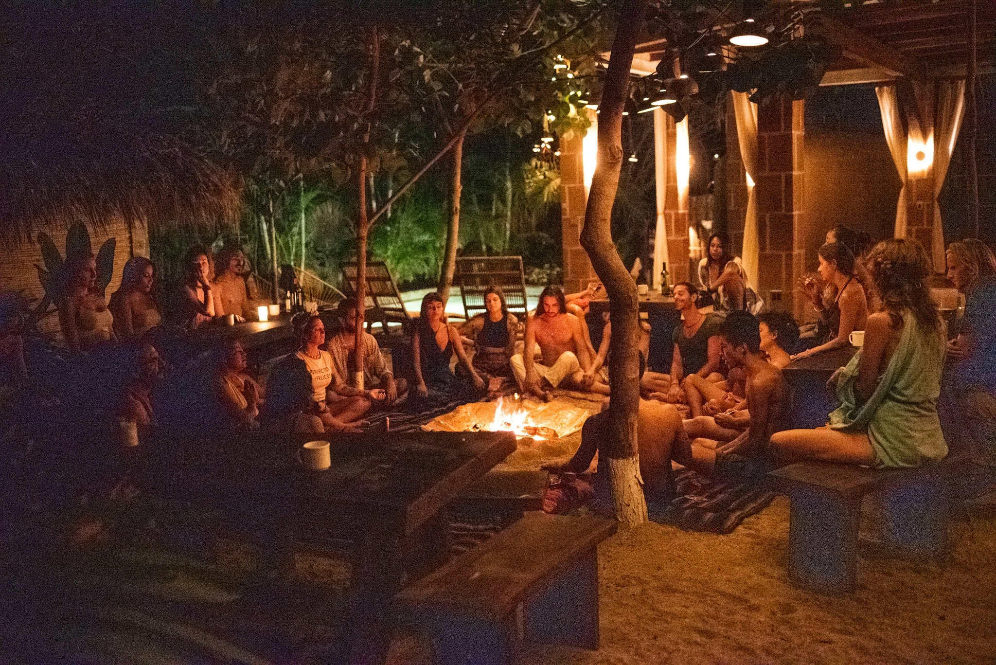 Cena nocturna con muchas personas sentadas alrededor de una fogata, en un entorno al aire libre con árboles, paredes de ladrillo y lámparas en las paredes.
