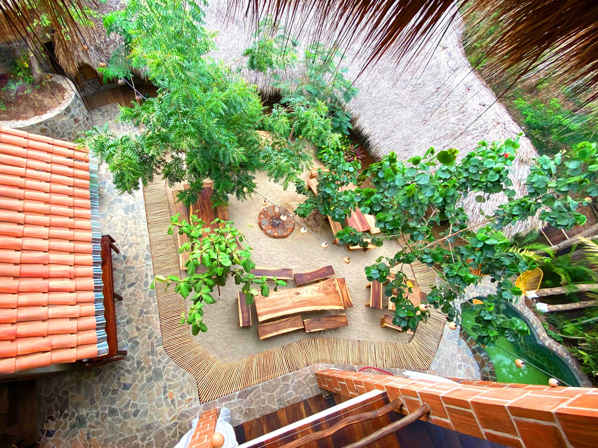 Vista aérea de un área de descanso con mesas de madera, plantas verdes, una estructura de techo de palapa y paredes de ladrillo, en un entorno natural.