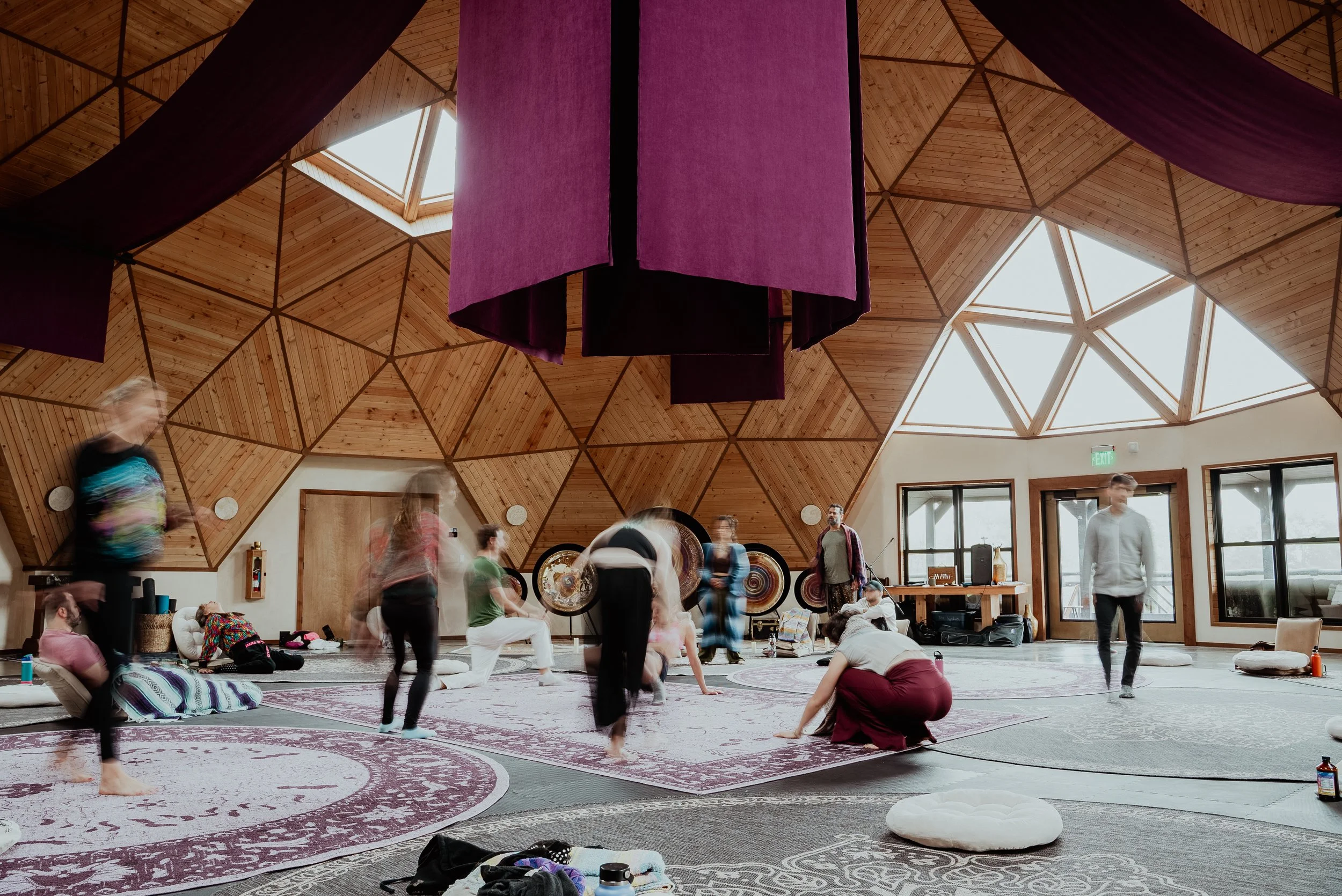 Interior de una sala de yoga con diversas personas participando en una clase, con paredes de madera y ventanas grandes que dejan entrar luz natural, con colchonetas y cojines en el piso.