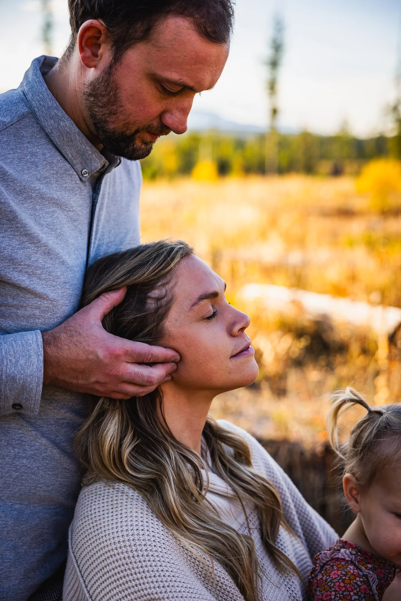 Dr. Peter Fennelly, a man with a beard and a woman with long wavy hair in an outdoor setting during autumn. The man gently holds the woman's head, feeling her TMJ to assess for motion.