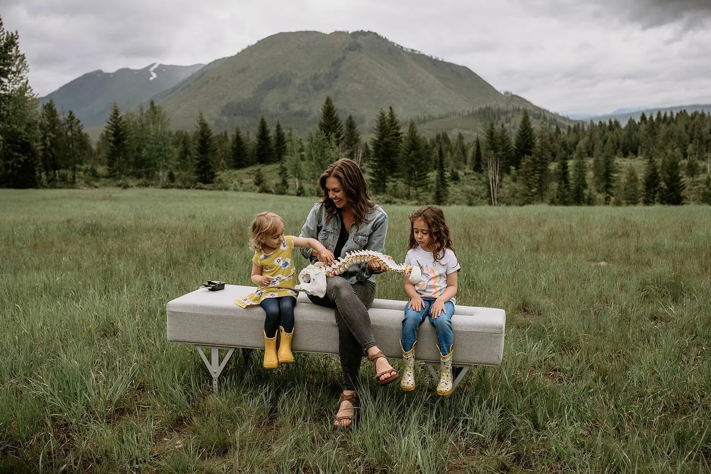 A woman with two young girls sitting on a gray bench outdoors in a grassy field with trees and mountains in the background. The woman holds a skeleton of a vertebrate, and the girls are reaching towards it, all appearing to be engaged and smiling.