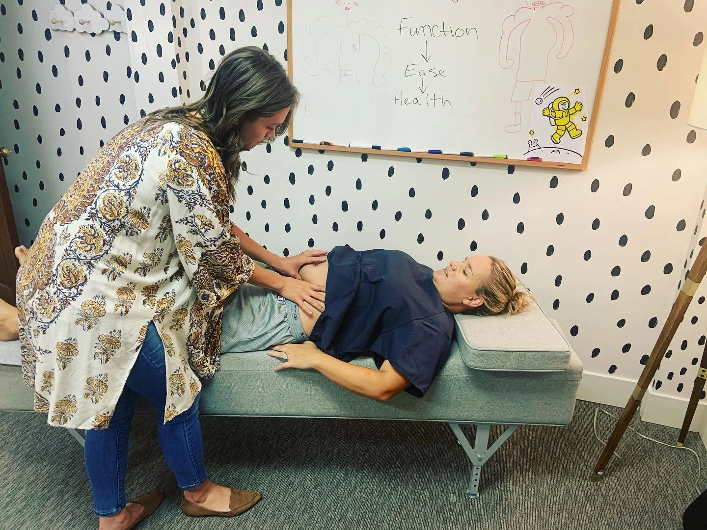 A healthcare worker performs an abdominal examination on a woman lying on an examination table in a clinical setting. The woman appears relaxed, and the healthcare worker is focused on the procedure. Behind them, there's a whiteboard with drawings and notes, including the words "Function," "Ease," and "Health." The room has patterned wallpaper with black dots and a small decorative cloud.