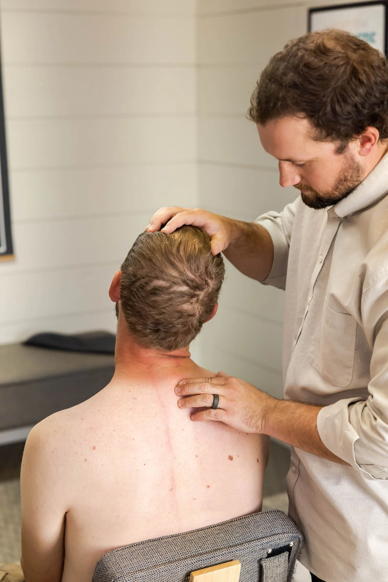 A man with short, light brown hair is being touched by another man with darker, curly hair on his head. The man with curly hair is leaning forward with his eyes closed, gently pressing his forehead to the top of the other man's head. The man with light brown hair is sitting shirtless, with his back to the camera, and resting his hand on a gray chair with a wooden handle.