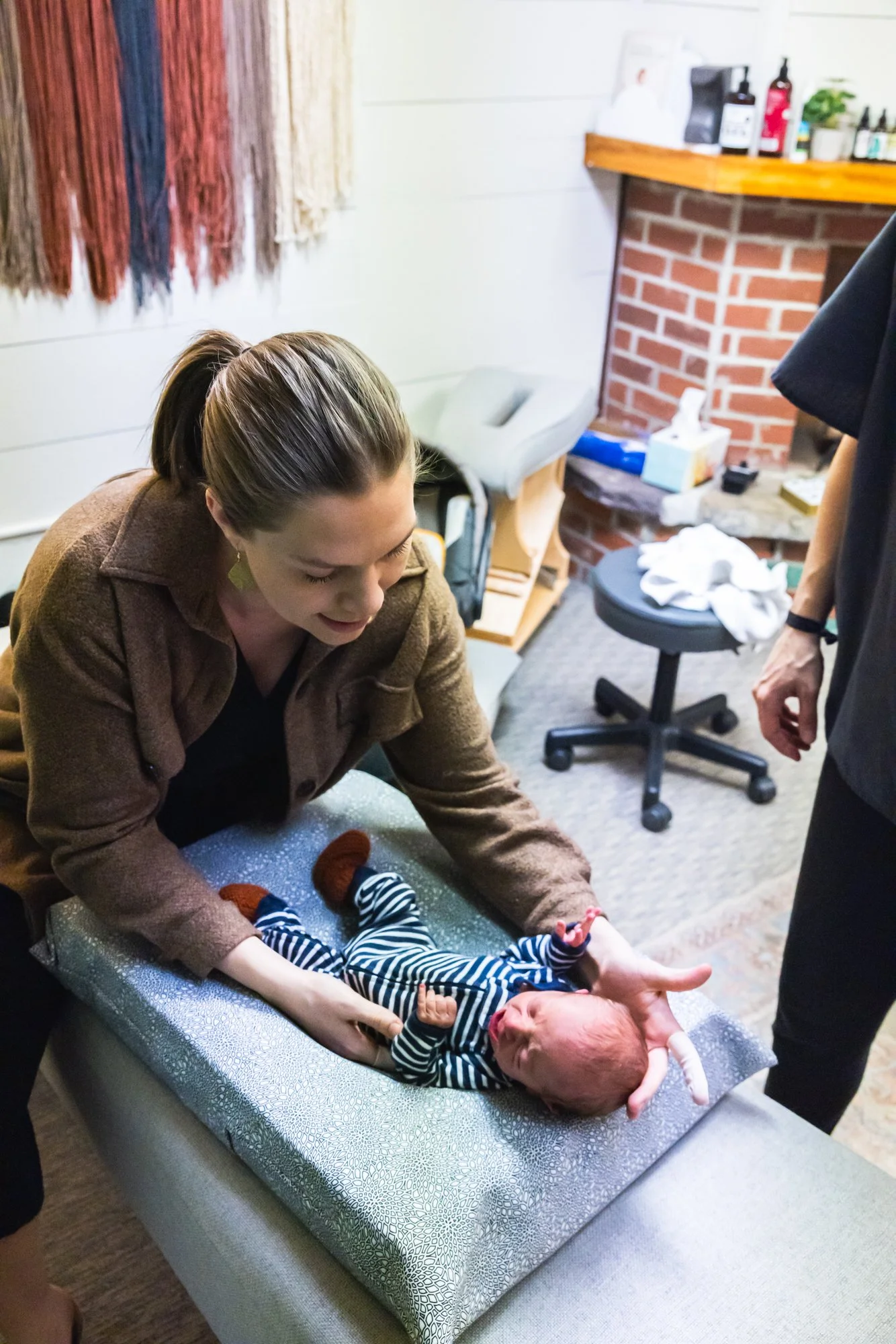 A woman lying on a table holding a newborn baby, while Dr. Jordan Fennelly of Two Sparrows Family Chiropractic assesses the newborn's spine with her chiropractic assessment.