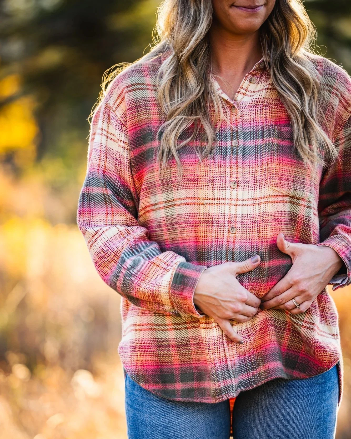 A woman wearing a red, orange, and gray plaid shirt with long, wavy blonde hair, standing outdoors with autumn-leafed trees in the background, gently holding her stomach with both hands.
