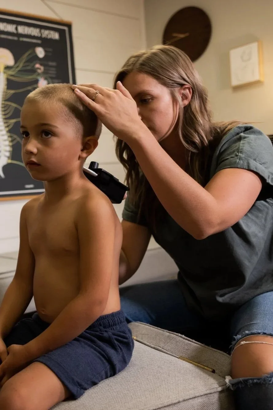 Dr. Jordan is checking a young boy's spinal temperature using a temperature differential device while the boy is sitting on a bench. The woman appears to be assessing his head with her hand to feel the motion of the joint.