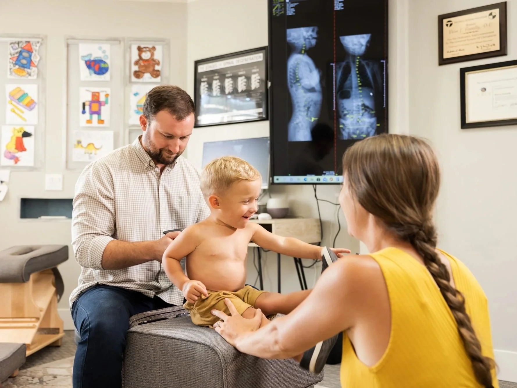A young boy sitting on an examination table at a doctor's office, smiling and reaching out while talking to a woman in a yellow sleeveless top. Dr. Pete stands beside him, looking down at the boy. The office has medical charts and X-rays on the wall.