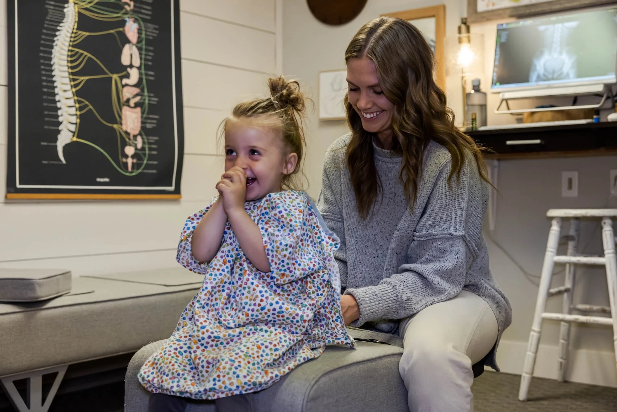 A young girl in a colorful hospital gown sitting on an examination table, smiling and covering her mouth with her hands while Dr Jordan Fennelly, sits behind her, smiling. They are in a chiropractic room with anatomy posters and a computer monitor.