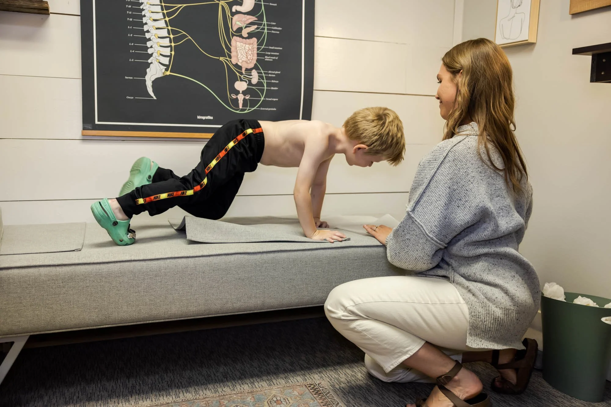 Dr. Jordan Fennelly is helping a boy lie down on the adjusting table in her office, Two Sparrows family Chiropractic