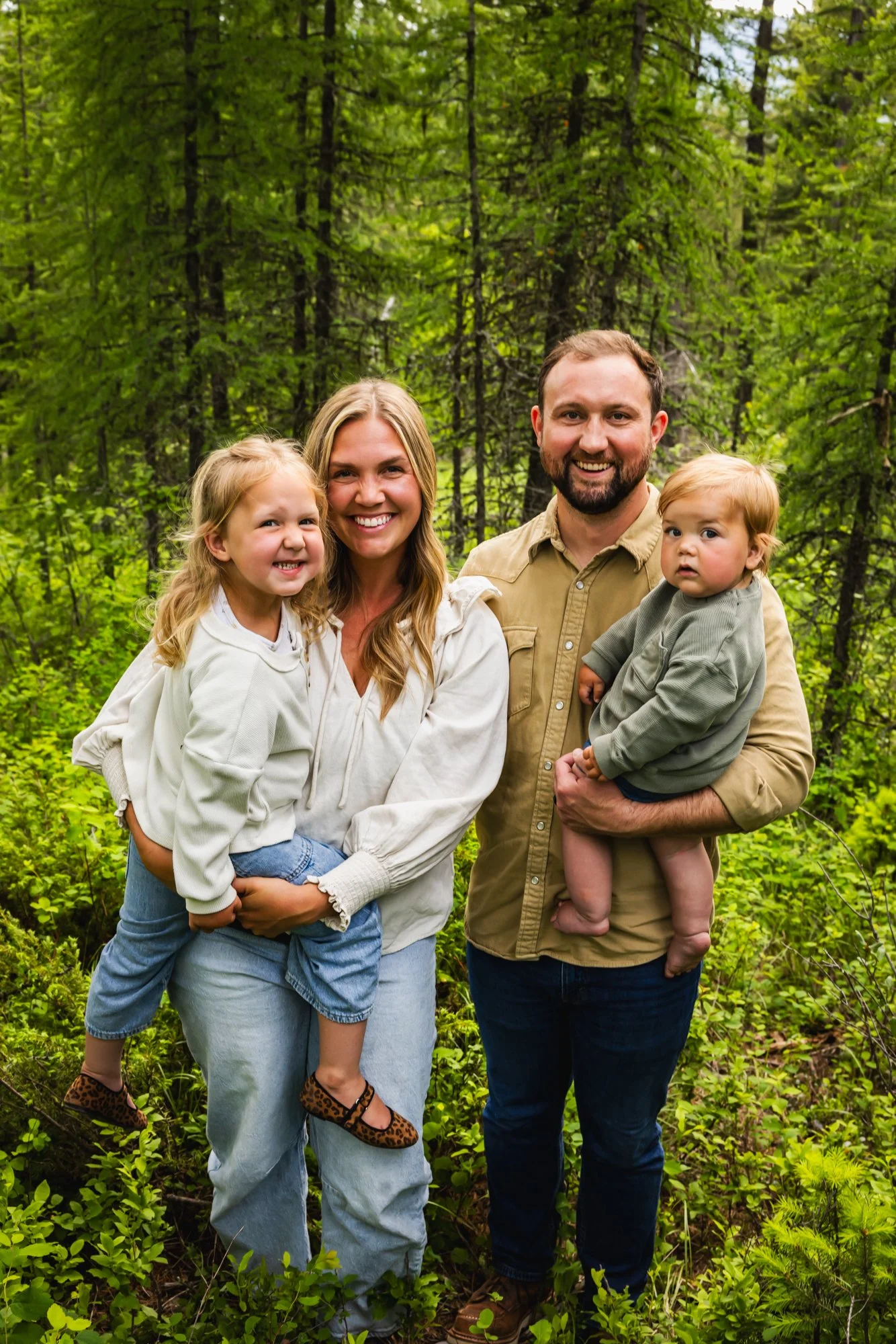 A family of four standing outdoors in a green forest, smiling at the camera.