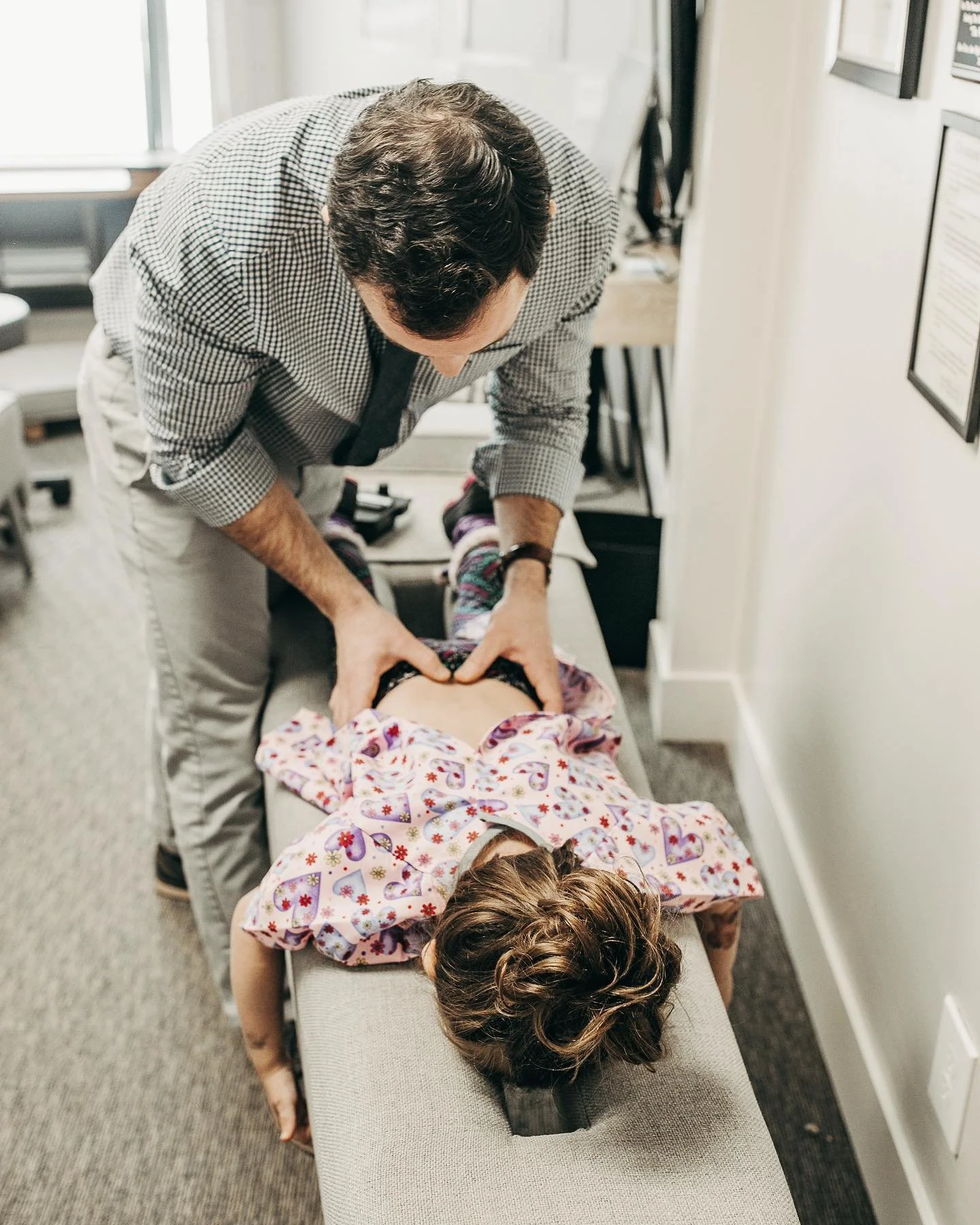 A chiropractor adjusts a young girl's back while she lies face down on an examination table in a medical office. The girl is wearing a hospital gown with colorful hearts and flowers, and the chiropractor is wearing a checkered shirt.