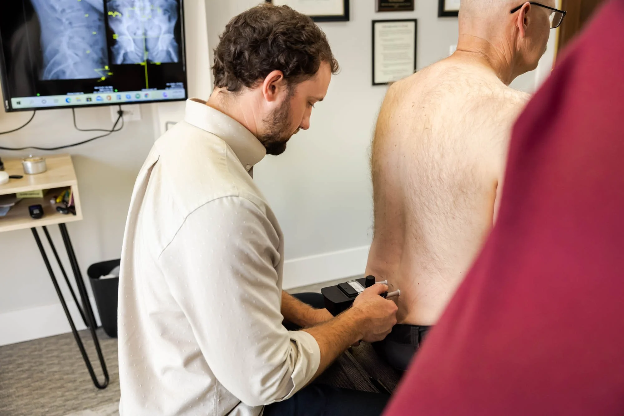A healthcare professional in a white coat is conducting a medical test on a male patient with a bare back. The professional is holding a medical device, likely a nerve conduction or electromyography instrument, on the patient's back. There is a television screen on the wall showing an X-ray or scan with green markers, and framed certificates are hung on the wall in the background.