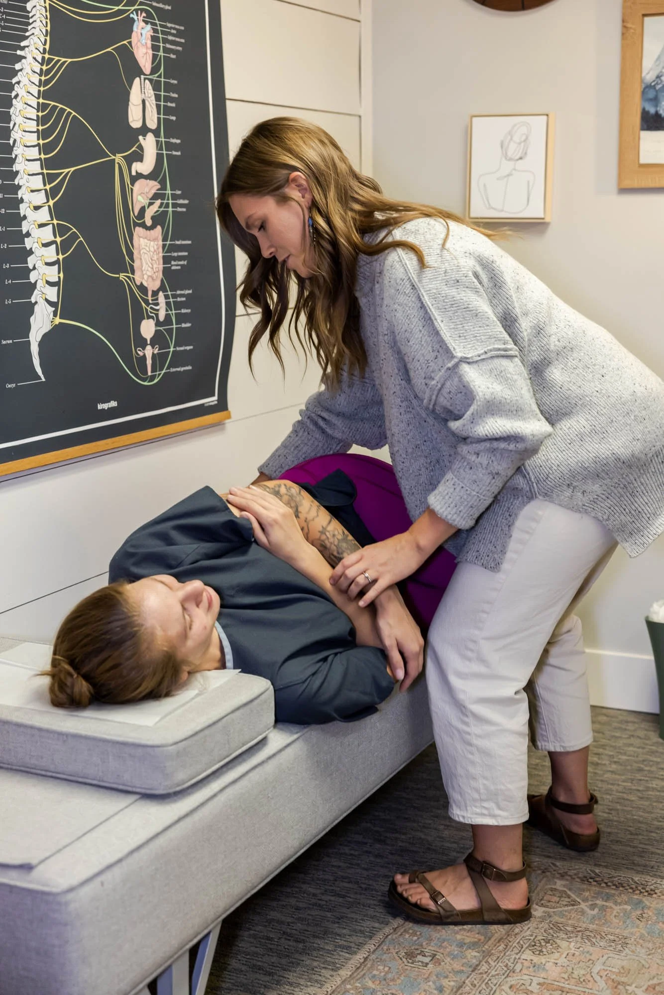 A woman performing a chiropractic adjustment on a woman lying on a pelvic bench. The setting appears to be a chiropractic office, Two Sparrows Family Chiropractic in Whitefish, MT, with anatomical charts on the wall.