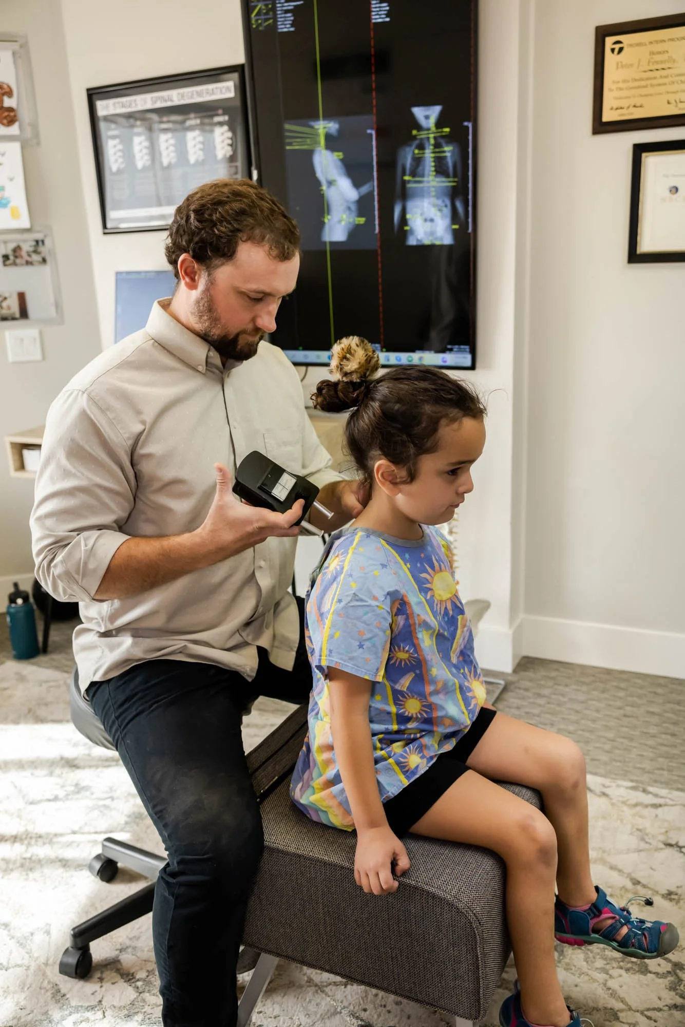 Dr. Peter Fennelly of Two Sparrows Family Chiropractic in Whitefish, Montana is in his office with a pediatric patient, with her x-rays on the screen, performing a chiropractic analysis of the patient's spine prior to performing an adjustment.