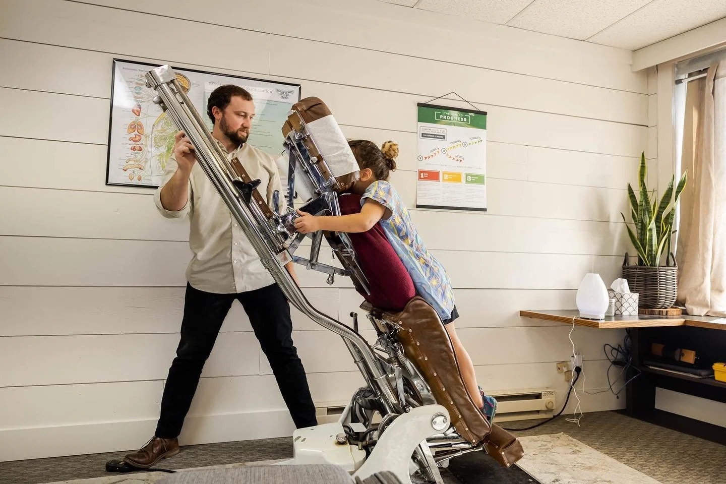 A girl in a colorful dress is sitting in a dental chair, leaning forward, with her head in a chiropractic headrest. Dr. Peter Fennelly is standing beside her adjusting the height of the chiropractic table before he adjusts her.