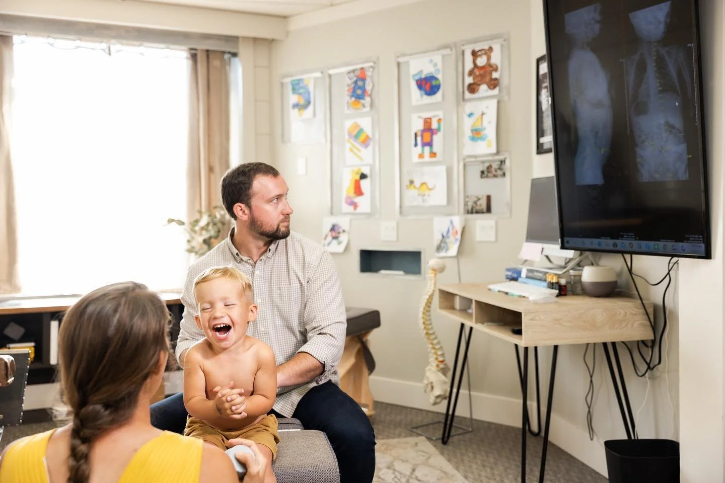 A family in a medical office, with a child laughing and a woman, possibly a parent, next to him. The man and woman are facing a television screen displaying an X-ray of a skull and spine. The room has colorful artwork on the wall, a human spine model, and medical supplies on a wooden stand.