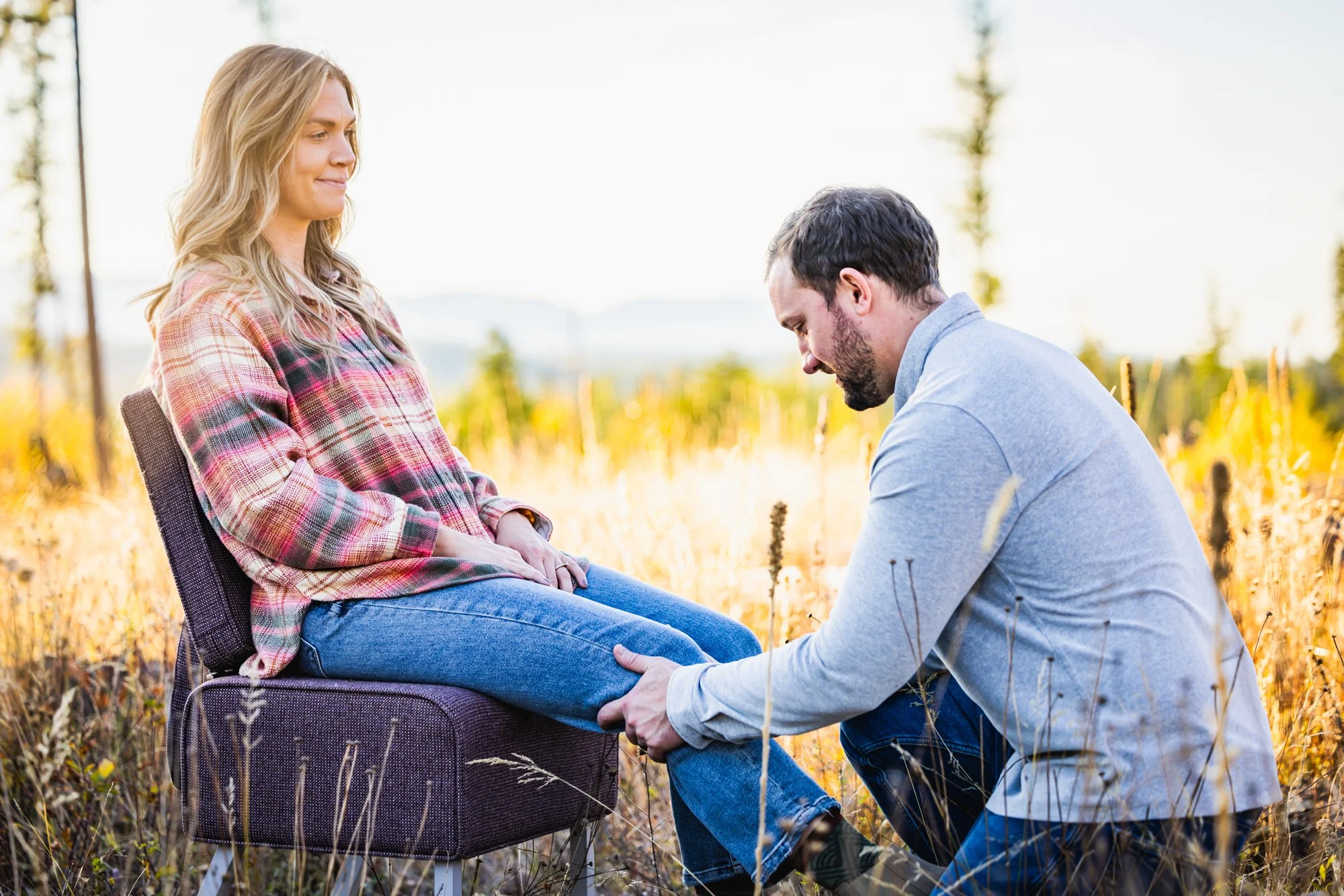 Dr. Peter Fennelly is assessing the knee joint of a patient. The woman is wearing a plaid shirt and jeans, and the man is wearing a gray shirt and jeans. They are surrounded by tall grass with trees in the background.
