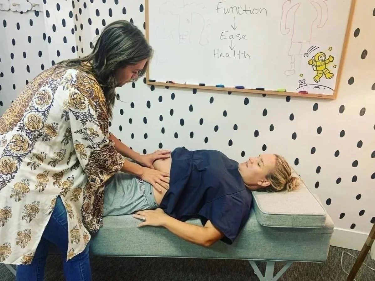 A pregnant woman lying on a chiropactic examination table while Dr. Jordan Fennelly, palpates her round ligament of her belly, during a consultation in a room with polka dot wall decor and a whiteboard with diagrams and notes.