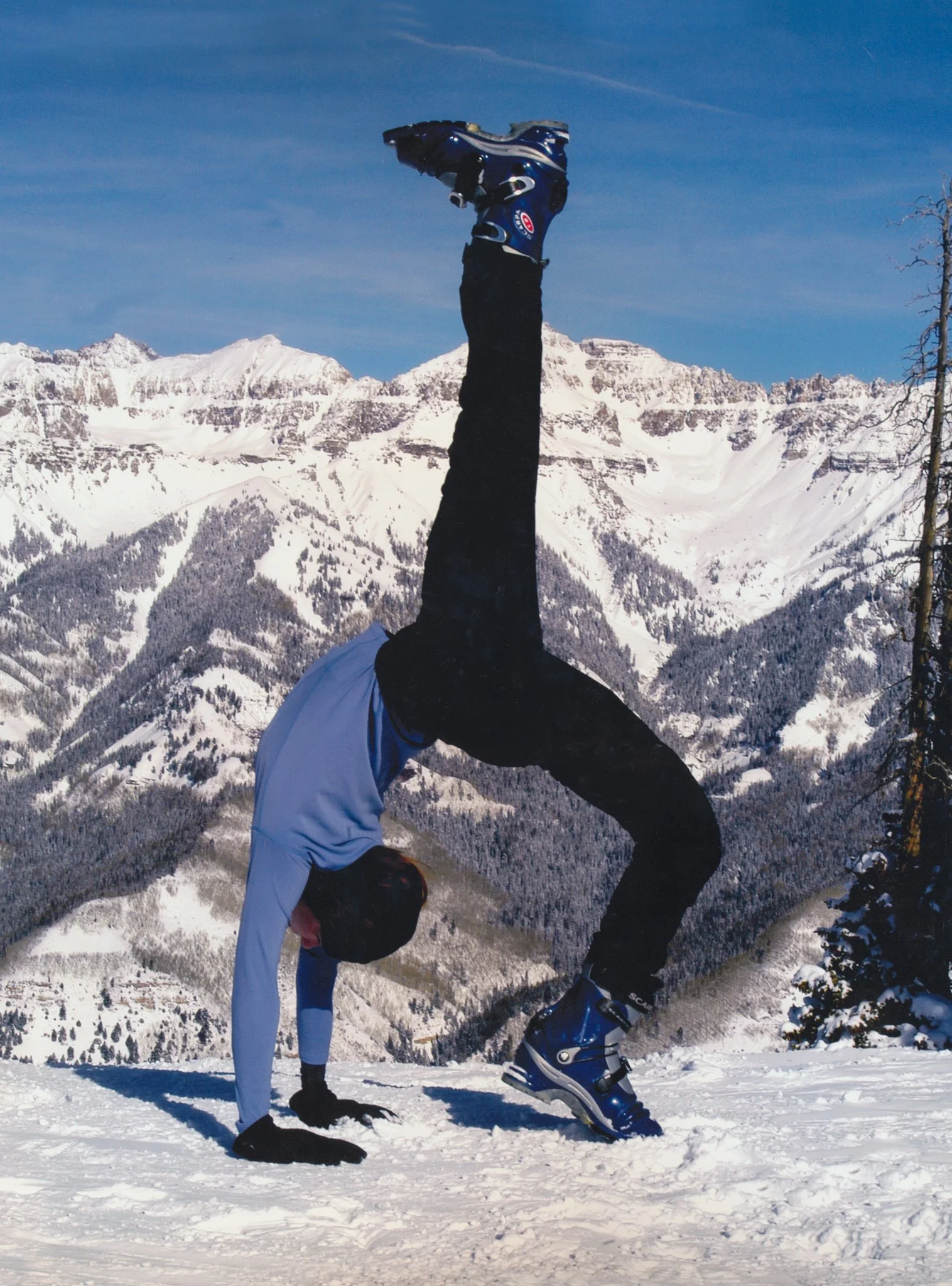 A person dressed in winter clothing performing a handstand on snow in a mountainous, snowy landscape.