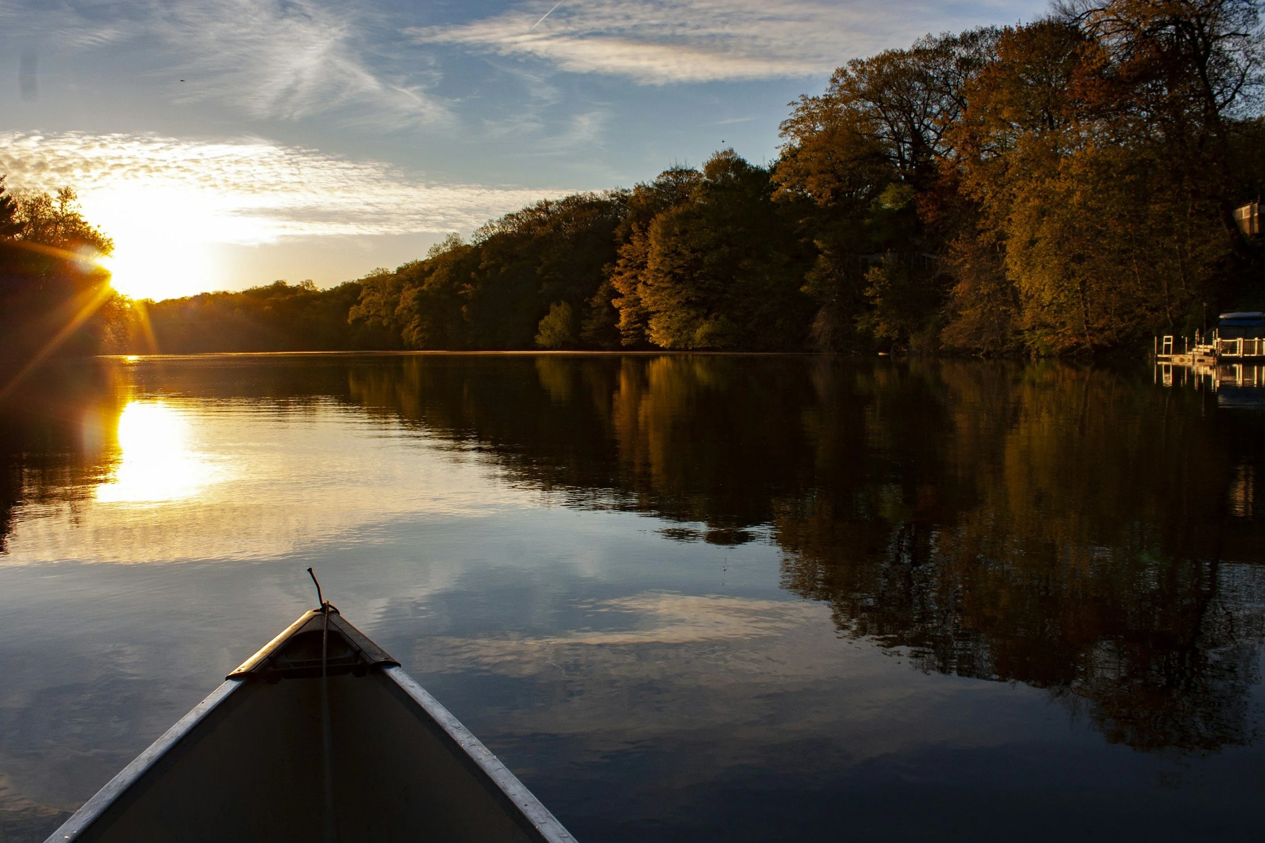 View from a canoe on a calm river with trees in autumn colors on the right and a sunset on the left side of the sky.