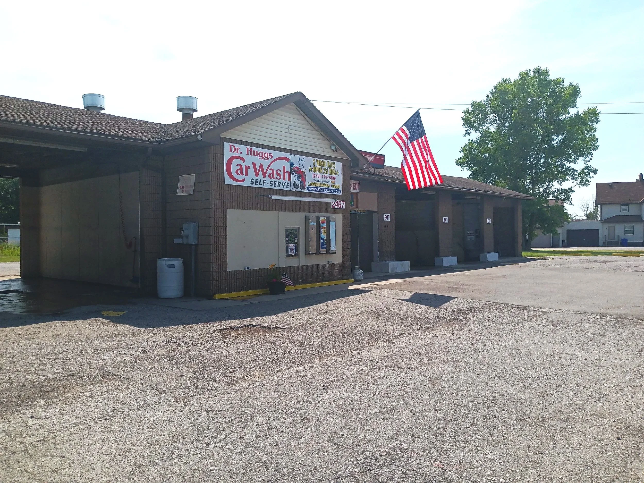 American flag flying outside a car wash business called Dr. Huggs Car Wash, with a gravel parking lot and a large green tree nearby.