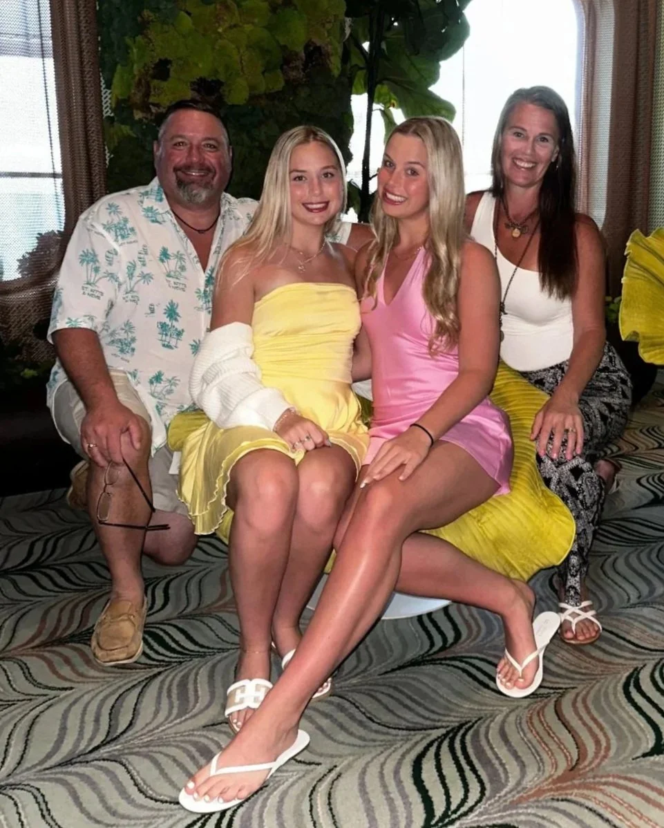 Four people sitting on a couch, three women and one man, smiling at the camera. The women are wearing pink, yellow, and white dresses; the man is wearing a Hawaiian shirt. The background features greenery and large windows.