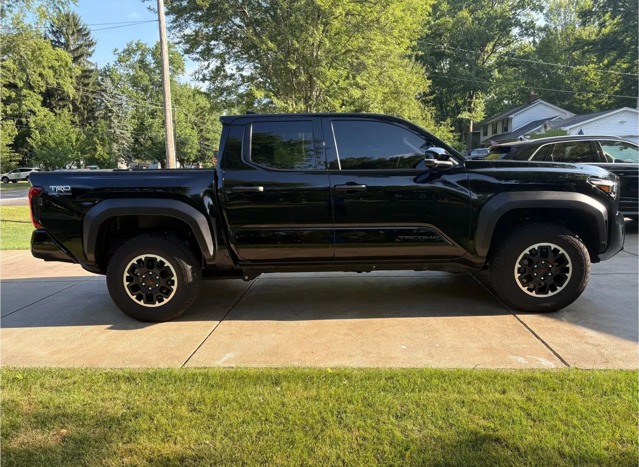 A black Toyota Tacoma pickup truck parked on a concrete driveway with a grassy lawn in front and a residential neighborhood background, including trees, houses, and parked cars.