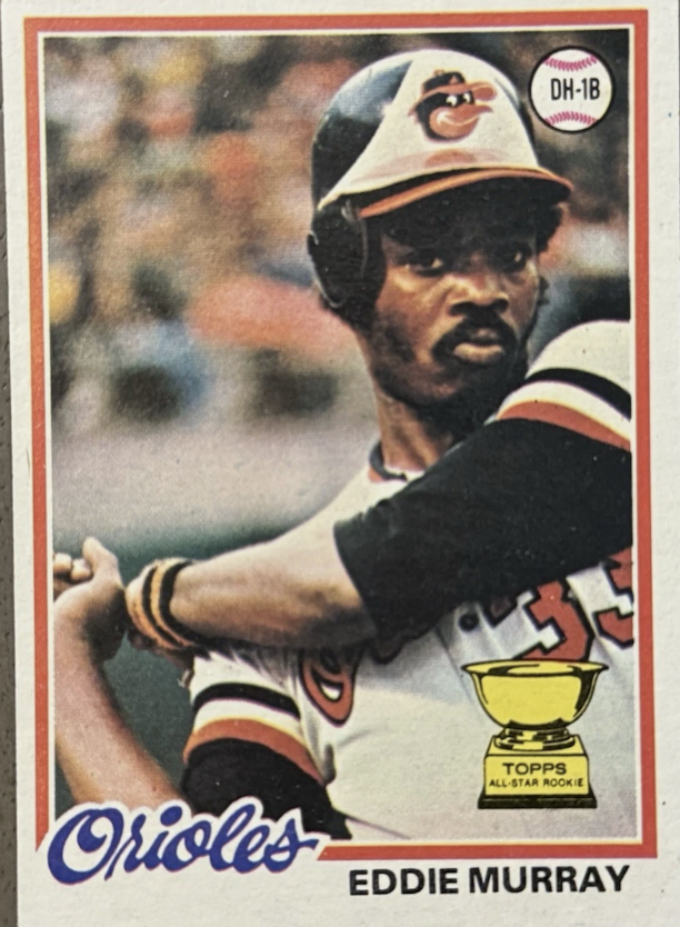 A baseball card featuring Eddie Murray of the Baltimore Orioles. He is wearing a baseball helmet with an Orioles logo and a uniform with the team name and number. The card has a Topps All-Star Rookie trophy illustration and the label DH-1B in the top right corner.