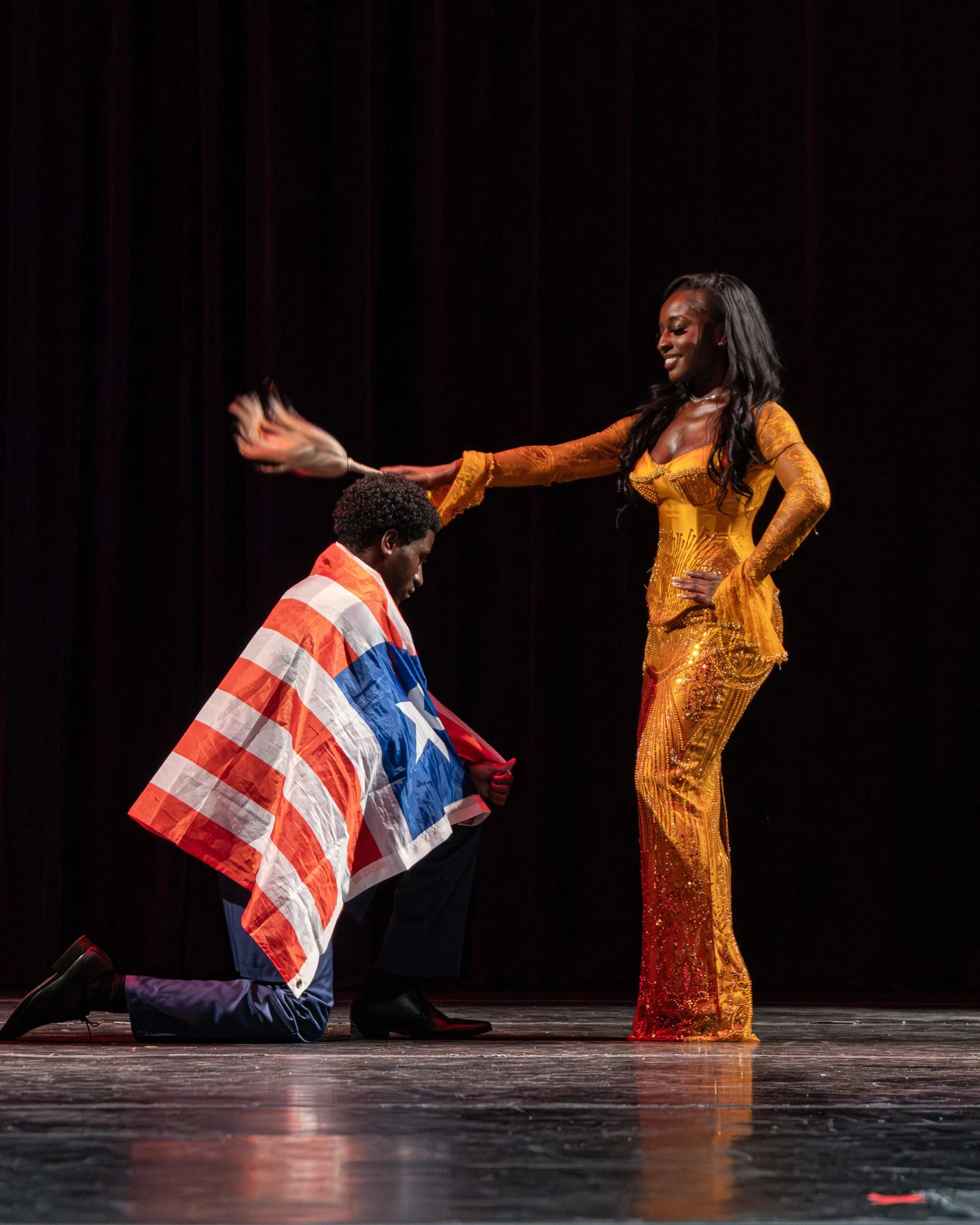 A woman dressed in a glittering gold gown standing on stage with a smile, while a man kneeling with his head bowed, holding a flag of the Netherlands.