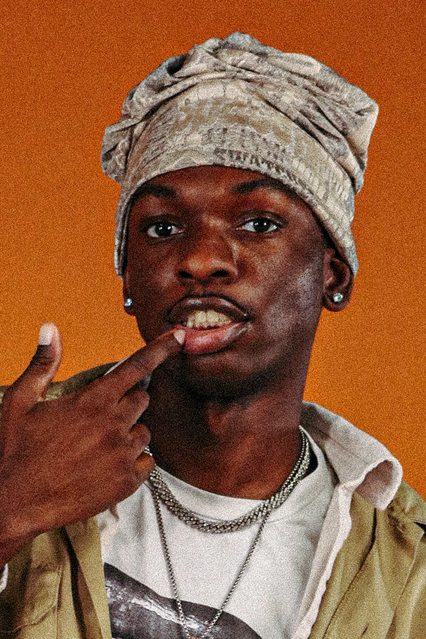 Close-up of a person with dark skin wearing a head wrap, jewelry, a graphic t-shirt, and a layered jacket, against an orange background.